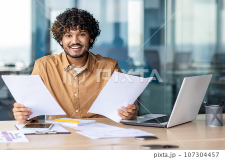 Portrait of young successful financier businessman paper work, hispanic smiling and looking at camera man holding paper reports and bills in hands, satisfied with achievement and checking results. Portrait of young successful financier businessman paper work, hispanic smiling and looking at camera man holding paper reports and bills in hands, satisfied with achievement and checking results. 107304457