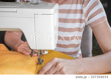Tailor hands stitching yellow fabric on modern sewing machine at workplace in atelier Tailor hands stitching yellow fabric on modern sewing machine at workplace in atelier 107308654