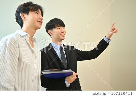 Asian male realtor wearing suit showing new house to smiling buyer. Real estate, loan and property investment concept Asian male realtor wearing suit showing new house to smiling buyer. Real estate, loan and property investment concept 107310411
