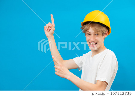 Happy handsome teen builder in a yellow helmet is looking at the camera and points fingers up, standing in a studio on a blue background 107312739