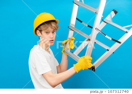 Young repairman teen in white t-shirt and gloves with yellow helmet in head, holding ladder in hands standing in studio on blue background. A future architect isolated on a blue background Young repairman teen in white t-shirt and gloves with yellow helmet in head, holding ladder in hands standing in studio on blue background. A future architect isolated on a blue background 107312740