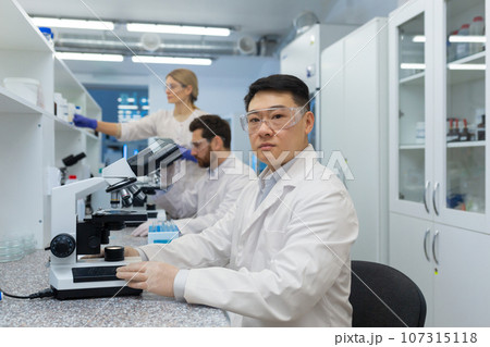 A group of young medical students and scientists are studying in the laboratory of the university. Asian young man sits behind a microscope and looks seriously at the camera. 107315118