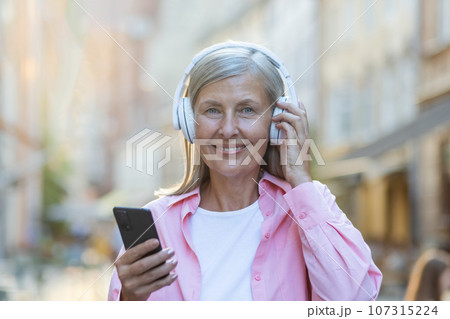 Portrait of an older beautiful gray-haired woman wearing headphones listening to music on the phone. Standing on a city street and smiling at the camera. Close-up photo. Portrait of an older beautiful gray-haired woman wearing headphones listening to music on the phone. Standing on a city street and smiling at the camera. Close-up photo. 107315224