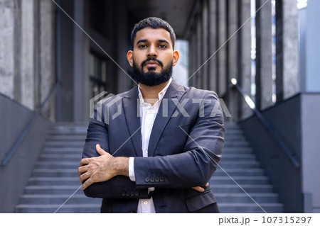 Portrait of a young Muslim male lawyer standing in a business suit outside a courthouse and looking confidently into the camera with his arms crossed. Portrait of a young Muslim male lawyer standing in a business suit outside a courthouse and looking confidently into the camera with his arms crossed. 107315297