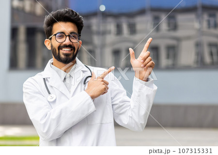Outdoor portrait of a young successful indian doctor outside the clinic, the doctor in a medical white coat is smiling and looking at the camera, pointing with his fingers in the direction backwards. Outdoor portrait of a young successful indian doctor outside the clinic, the doctor in a medical white coat is smiling and looking at the camera, pointing with his fingers in the direction backwards. 107315331