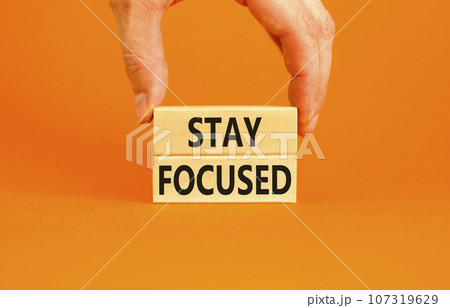Stay focused symbol. Concept words Stay focused on wooden blocks on a beautiful orange background. Businessman hand. Business, support, motivation, psychological and stay focused concept. Copy space. Stay focused symbol. Concept words Stay focused on wooden blocks on a beautiful orange background. Businessman hand. Business, support, motivation, psychological and stay focused concept. Copy space. 107319629