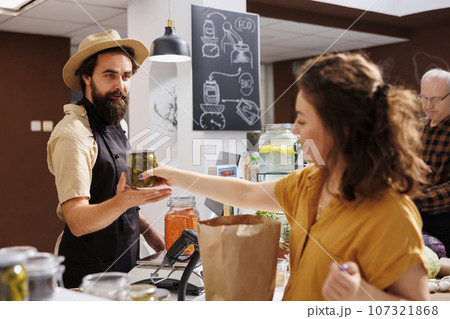 Cheerful merchant at checkout counter in zero waste shop selling vegan products to environmentally conscious client. Woman purchasing healthy food in low carbon footprint local neighborhood store 107321868