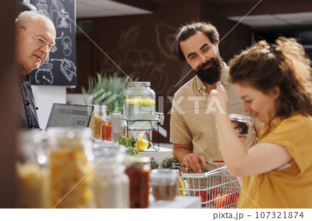 Husband and wife at zero waste supermarket checkout counter with shopping basket full of organic food. Customers waiting for their pantry staples to be weighted with electronic scale in local shop Husband and wife at zero waste supermarket checkout counter with shopping basket full of organic food. Customers waiting for their pantry staples to be weighted with electronic scale in local shop 107321874