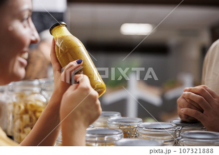 Vendor showcasing her products in zero waste marketplace venue, selling homemade fruit juice to interested customer, close up. Local farmer trading healthy food items during event, blurry background 107321888