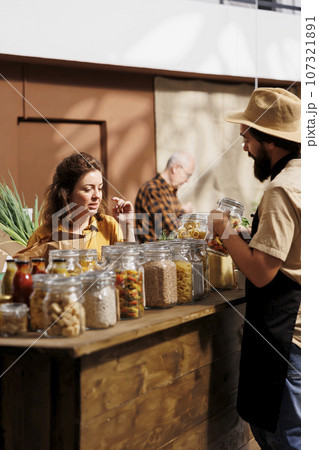 Client shopping for pantry staples in zero waste store using plastic free paper bag, being assisted with information by trader. Seller in no single use plastics policy shop showing woman products 107321891