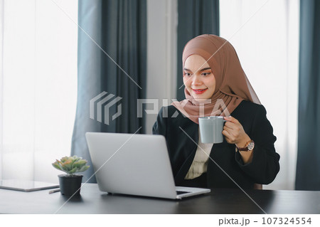 Smiling happy arab asian muslim woman in brown hijab working with laptop computer at modern office. 107324554