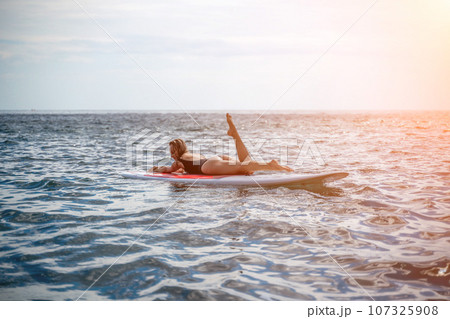 Woman sup sea. Sports girl on a surfboard in the sea on a sunny summer day. In a black bathing suit, he sits on a sapa in the sea. Rest on the sea. Woman sup sea. Sports girl on a surfboard in the sea on a sunny summer day. In a black bathing suit, he sits on a sapa in the sea. Rest on the sea. 107325908
