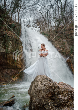 Happy woman in a white dress stands on a stone with a waterfall behind 107325915