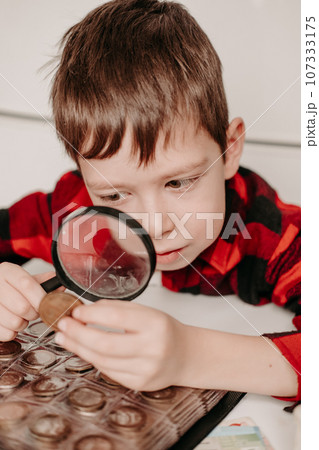 collecting coins. The boy examines an old coin through a magnifying glass. Dollars, euros on the table. Money of different nations. Savings in the form of currencies and coins collecting coins. The boy examines an old coin through a magnifying glass. Dollars, euros on the table. Money of different nations. Savings in the form of currencies and coins 107333175