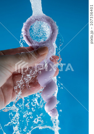 Woman holding lilac anal beads under running water on blue background. Sex toy hygiene concept. Woman holding lilac anal beads under running water on blue background. Sex toy hygiene concept. 107336401