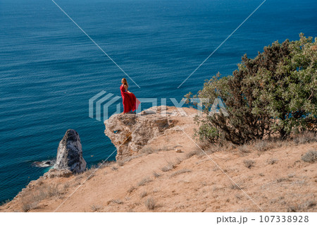 A woman in a red flying dress fluttering in the wind, against the backdrop of the sea. A woman in a red flying dress fluttering in the wind, against the backdrop of the sea. 107338928
