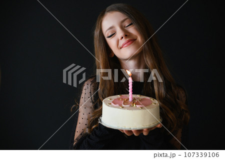 Happy teen girl wearing black dress holding birthday cake. Black background. 107339106