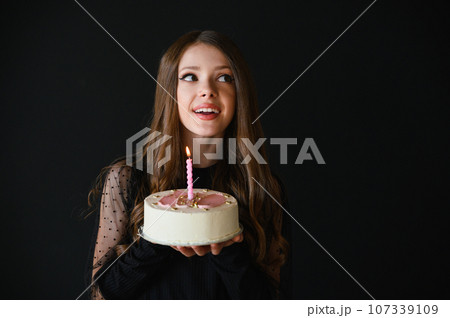 Happy teen girl wearing black dress holding birthday cake. Black background. 107339109