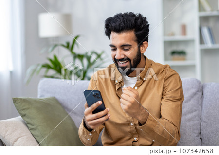 Young man received an online notification of a winning message sitting at home in the living room on the sofa, the winner is satisfied with results of the achievement joyfully holding his hand up. 107342658