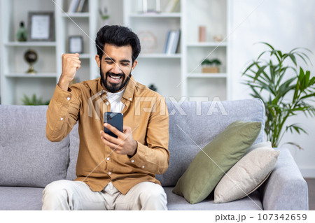 Young man received an online notification of a winning message sitting at home in the living room on the sofa, the winner is satisfied with results of the achievement joyfully holding his hand up. Young man received an online notification of a winning message sitting at home in the living room on the sofa, the winner is satisfied with results of the achievement joyfully holding his hand up. 107342659