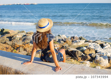 Beautiful girl in a hat sits near the sea on stones Beautiful girl in a hat sits near the sea on stones 107343994