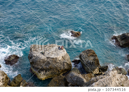MELLIEHA, MALTA - 01 JAN, 2020: A man sitting on a rock near the ocean on the island of Malta during calm sea MELLIEHA, MALTA - 01 JAN, 2020: A man sitting on a rock near the ocean on the island of Malta during calm sea 107344516