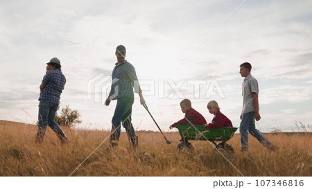 Mother carries baby and father pulls brothers in cart walking with family along field at sunset. Children with parents enjoy family weekend in farm field. Farmer family with children rest in field Mother carries baby and father pulls brothers in cart walking with family along field at sunset. Children with parents enjoy family weekend in farm field. Farmer family with children rest in field 107346816