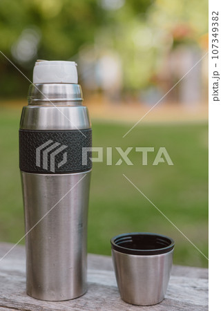 Stainless steel cup, mug with thermos stands on a wooden table in the forest on a green natural background. Thermos and mug with hot drink standing on wet wooden table after rain outdoors, close-up Stainless steel cup, mug with thermos stands on a wooden table in the forest on a green natural background. Thermos and mug with hot drink standing on wet wooden table after rain outdoors, close-up 107349382