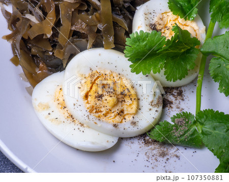 Seaweed salad and cut boiled egg. Dish on the table in a plate. Peppered half of an egg. Parsley greens as decoration. Seaweed salad and cut boiled egg. Dish on the table in a plate. Peppered half of an egg. Parsley greens as decoration. 107350881
