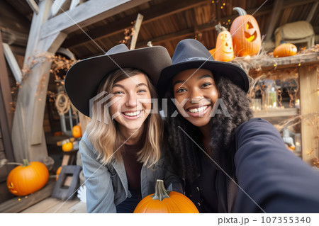 Two multiracial young women friends wearing witch hats having fun and take selfies at Halloween 107355340