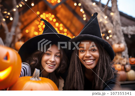 Two multiracial young women friends wearing witch hats having fun and take selfies at Halloween 107355341