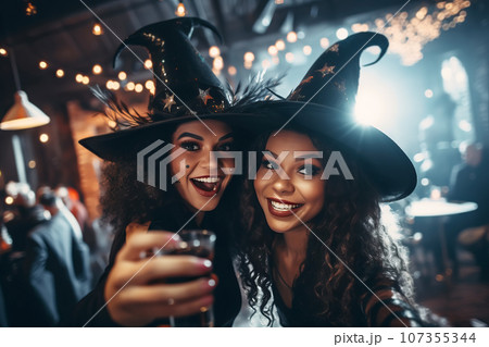 Two multiracial young women friends wearing witch hats having fun at Halloween party Two multiracial young women friends wearing witch hats having fun at Halloween party 107355344