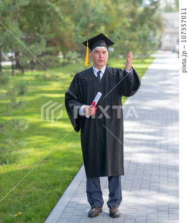 Serious old man in graduation gown holding diploma outdoors. 107357011