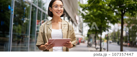 Portrait of young female student, standing near building on street, holding tablet and smiling 107357447