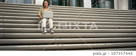Asian girl student sits on stairs near campus, types on laptop, does her homework outdoors 107357448