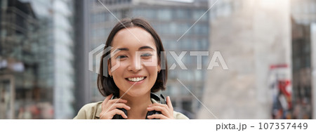 Vertical shot of beautiful asian woman posing with headphones around neck, smiling and laughing, standing on street in daylight Vertical shot of beautiful asian woman posing with headphones around neck, smiling and laughing, standing on street in daylight 107357449