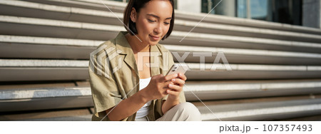 People and technology. Smiling beautiful asian woman sitting on stairs in city, holding mobile phone. Girl with smartphone rests outside 107357493