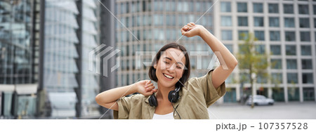 People. Portrait of smiling asian girl with headphones, dancing on street of city centre, looking happy People. Portrait of smiling asian girl with headphones, dancing on street of city centre, looking happy 107357528