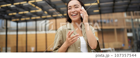 Young asian woman smiles, talks on mobile phone and walks in city centre, poses outdoors 107357544