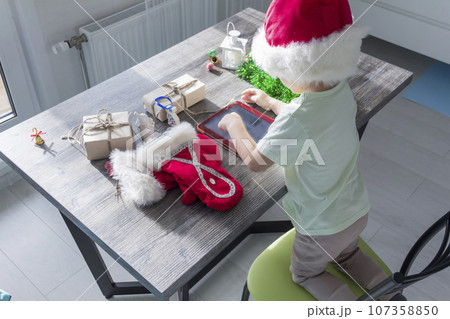 A small child in a Santa Claus hat, with Christmas gifts, is sitting at a New Year's table with a tablet in his hands. 107358850