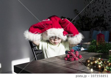 A small child in a Santa Claus hat and red mittens with Christmas gifts is sitting at the New Year's table. A small child in a Santa Claus hat and red mittens with Christmas gifts is sitting at the New Year's table. 107358851