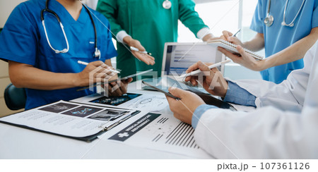 Medical team having a meeting with doctors in white lab coats and surgical scrubs seated at a table discussing a patients working online using computers in the medical industry.. 107361126