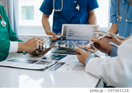 Medical team having a meeting with doctors in white lab coats and surgical scrubs seated at a table discussing a patients working online using computers in the medical industry.. 107361128