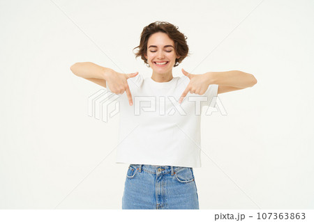 Image of beautiful, happy brunette woman, pointing fingers down, showing advertisement, standing over white studio background 107363863