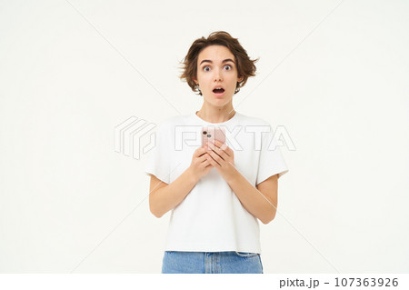 Portrait of brunette girl with shocked face, reading something amazing on mobile phone, standing with smartphone over white background Portrait of brunette girl with shocked face, reading something amazing on mobile phone, standing with smartphone over white background 107363926
