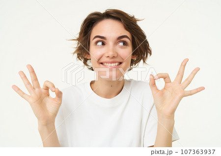 Image of excited brunette woman, smiles, shows okay, ok gesture, looks amused and happy, stands over white studio background 107363937