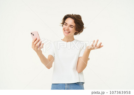 Potrait of woman video chats with friend, showing around, demonstrating something, has an online meeting on smartphone, standing over white background Potrait of woman video chats with friend, showing around, demonstrating something, has an online meeting on smartphone, standing over white background 107363958