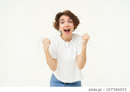 Cheerful brunette woman winning, triumphing, celebrating victory, achieves goal, stands over white background Cheerful brunette woman winning, triumphing, celebrating victory, achieves goal, stands over white background 107366055