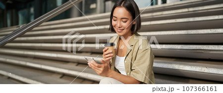 Happy urban girl drinks her takeaway coffee and scrolls feed on smartphone. Asian woman sits on stairs with tea and holding mobile phone 107366161