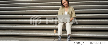 Portrait of young asian woman using her laptop, sitting outdoors on stairs. Happy smiling girl with personal computer 107366162